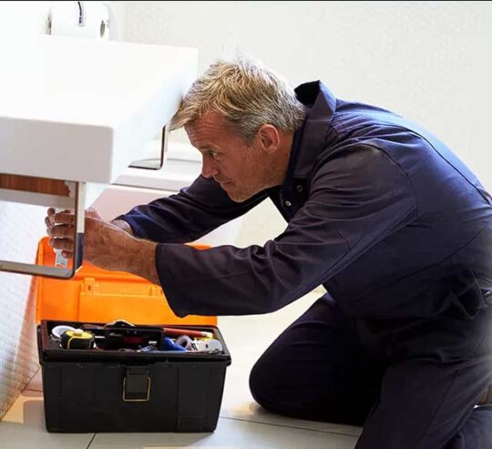 A plumber on the floor while fixing pipes under a white bathroom sink